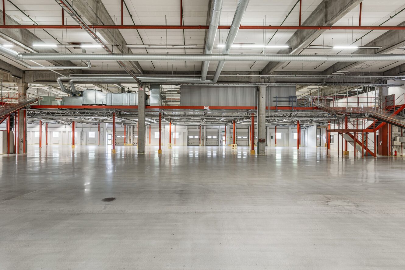 Stort, tomt industrilager med betonggolv, röda bärande balkar, synliga tak och trappor som leder till mezzaninvåningar på båda sidor.