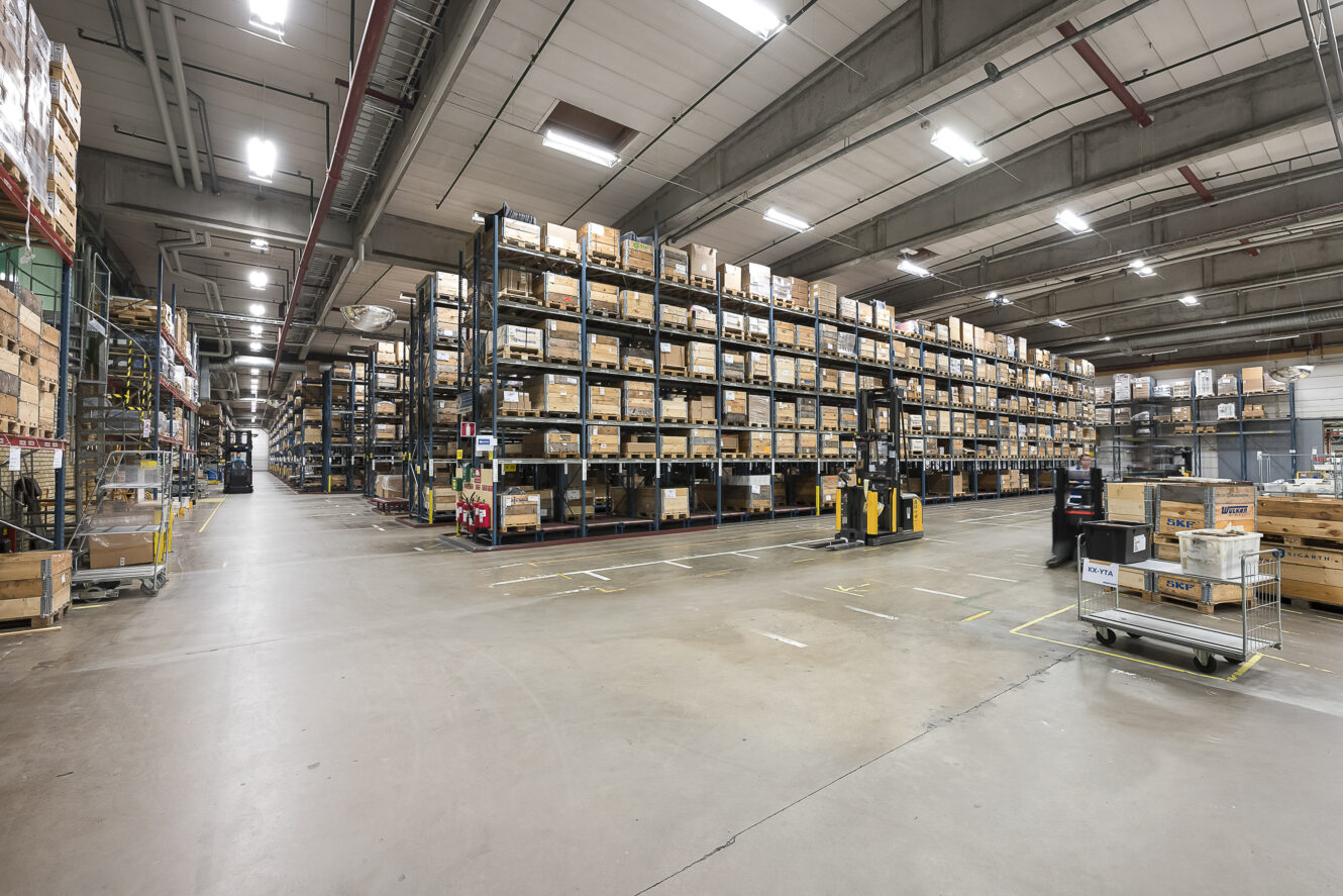 Stor lagerlokal med höga hyllor fyllda med lådor, gaffeltruckar och vagnar på golvet, under stark takbelysning.
