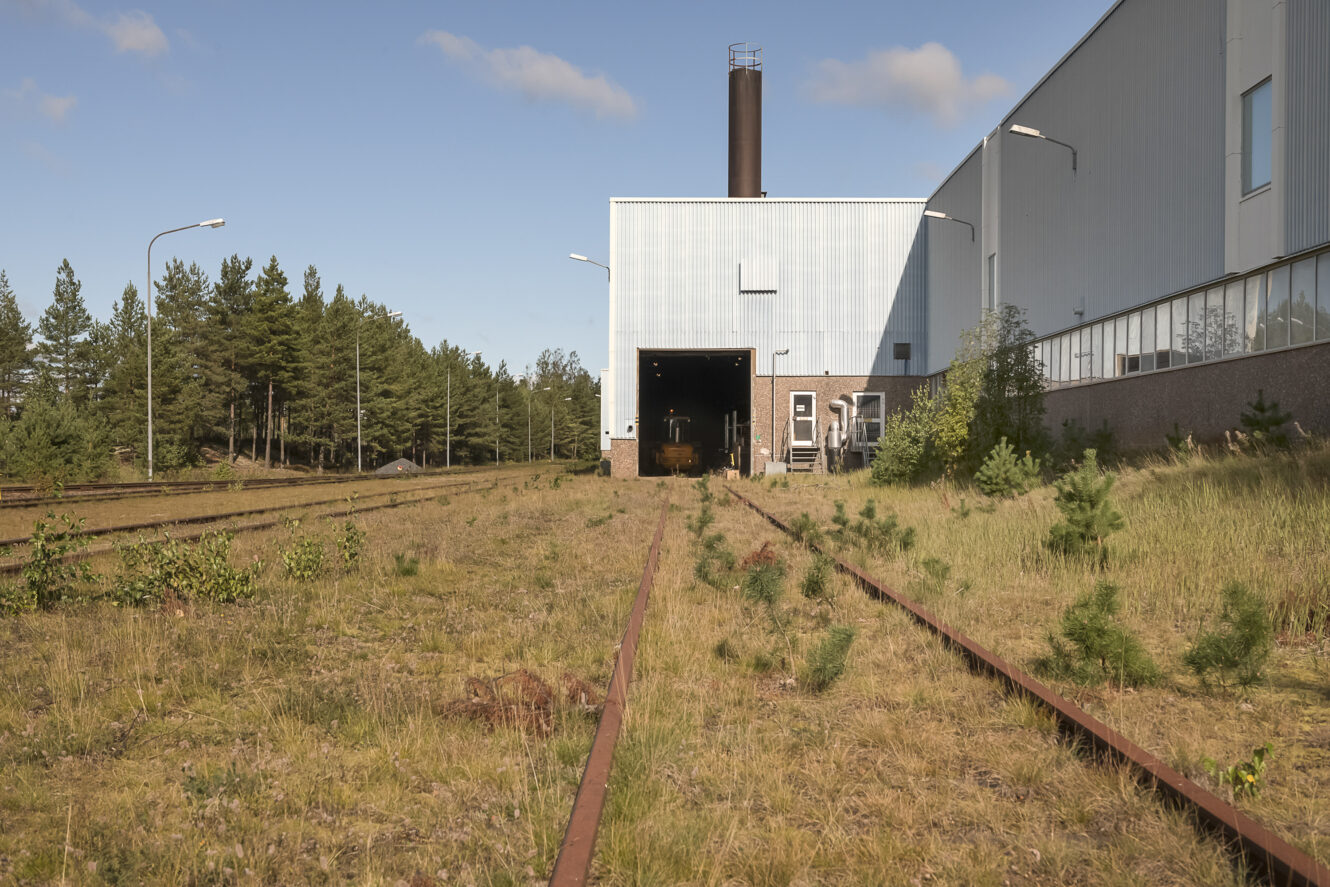 Nedlagda järnvägsspår som leder till en stor industribyggnad omgiven av övervuxet gräs och små träd, med en skog i bakgrunden.