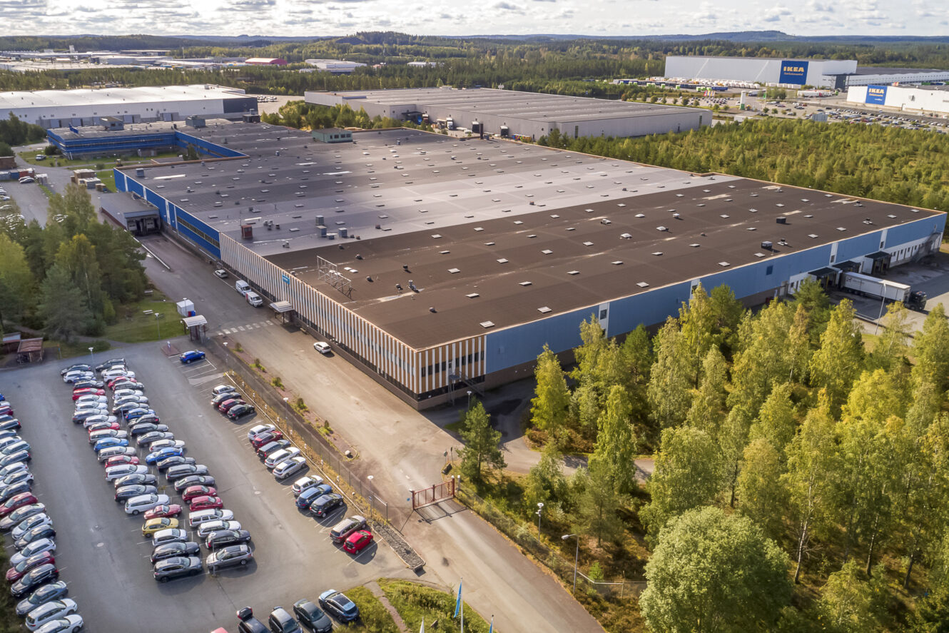 Flygfoto över en stor industrihall med en parkeringsplats full av bilar, omgiven av träd och andra industribyggnader.