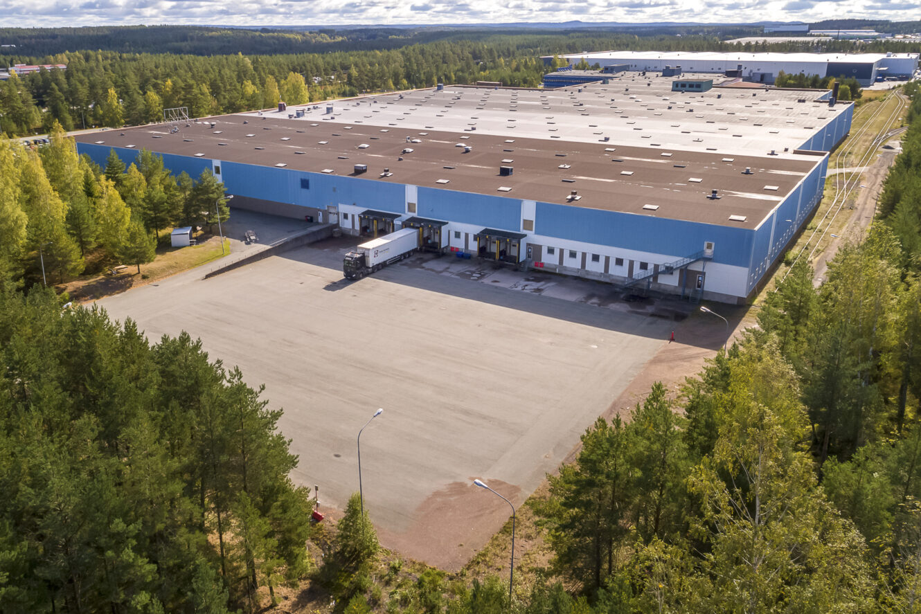 En stor blåvit lagerlokal med flera lastkajer, lastbilar parkerade utanför och omgiven av träd under en delvis molnig himmel.