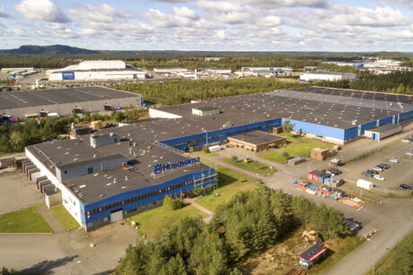 Flygfoto över ett stort industrikomplex med blå takbyggnader märkta Husqvarna, omgivet av vägar, parkeringsplatser och grönska under en delvis molnig himmel.