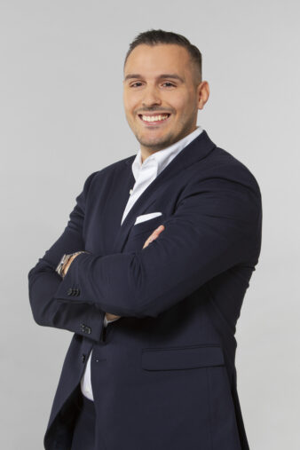 En man i mörk kostym och vit skjorta står med armarna i kors och ler mot kameran mot en vanlig ljus bakgrund.