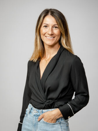 En kvinna med rakt axellångt hår, klädd i svart långärmad blus och ljusblå jeans, står och ler mot en grå bakgrund.