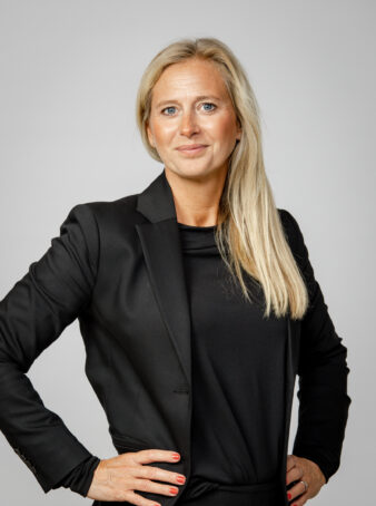 En kvinna med långt blont hår, klädd i svart kavaj och svart topp, står med händerna på höfterna mot en grå bakgrund.