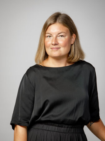 En kvinna med rakt, axellångt blont hår, klädd i en lös svart topp, står mot en ljusgrå bakgrund och tittar in i kameran med ett neutralt uttryck.