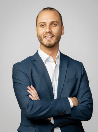 Man i blå kostym och vit skjorta står med armarna i kors och ler mot en ljusgrå bakgrund.