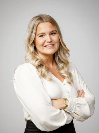 En kvinna med långt blont hår, klädd i vit blus och svarta byxor, står med armarna i kors och ler mot kameran mot en ljus bakgrund.