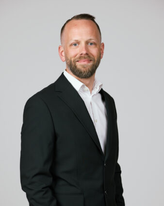 En man med skägg i svart kostym och vit skjorta står vänd framåt och ler mot en ljusgrå bakgrund.