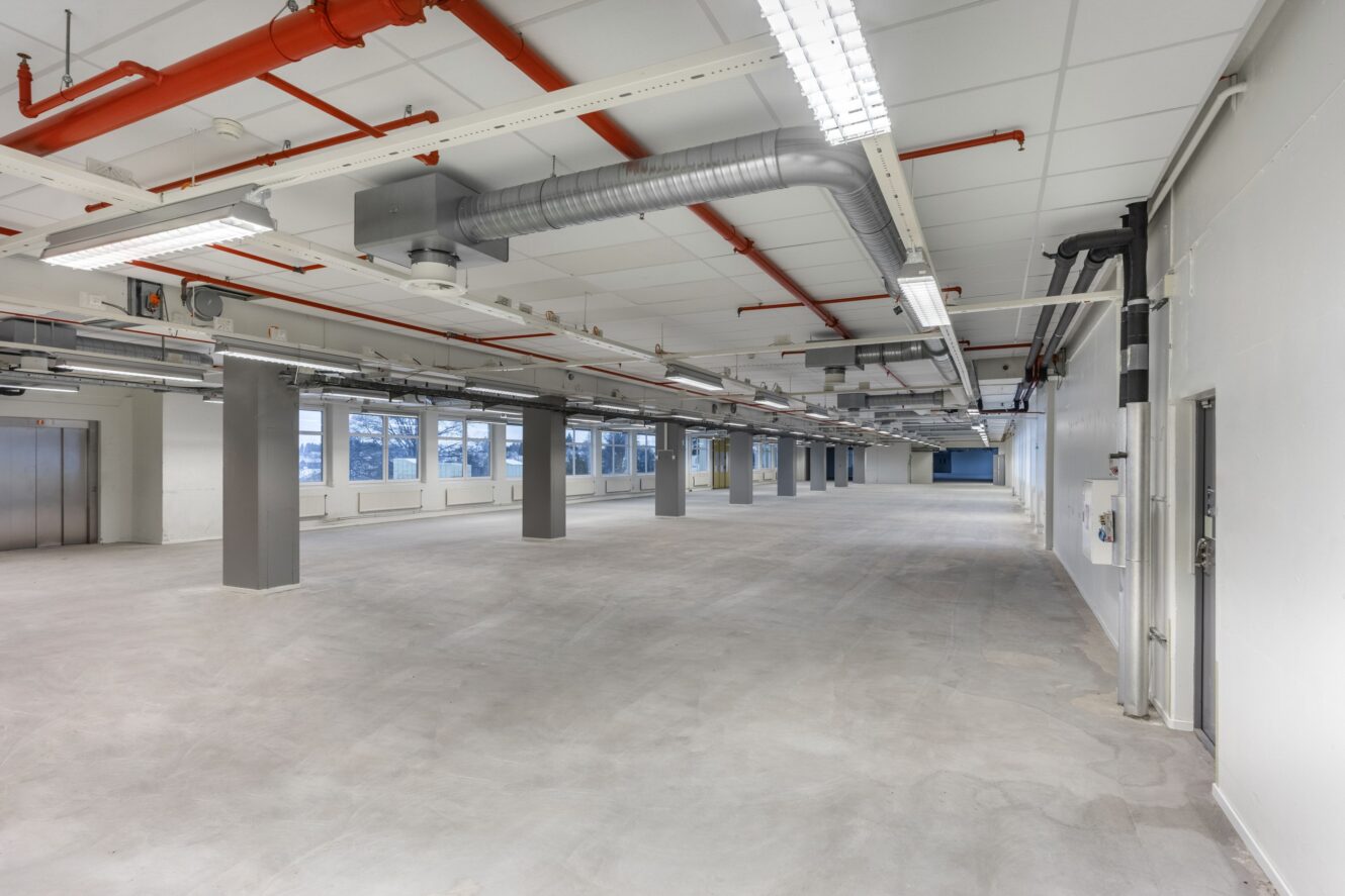 Ett stort, tomt industrirum med synliga rör, ventiler, pelare, lysrör i taket och flera fönster längs ena väggen.