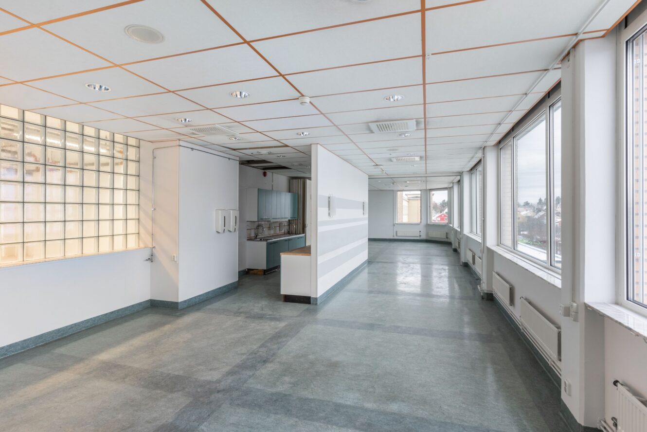 Ett stort, tomt rum med kaklat tak, stora fönster längs ena väggen och ett öppet pentry med skåp och bänkskivor.