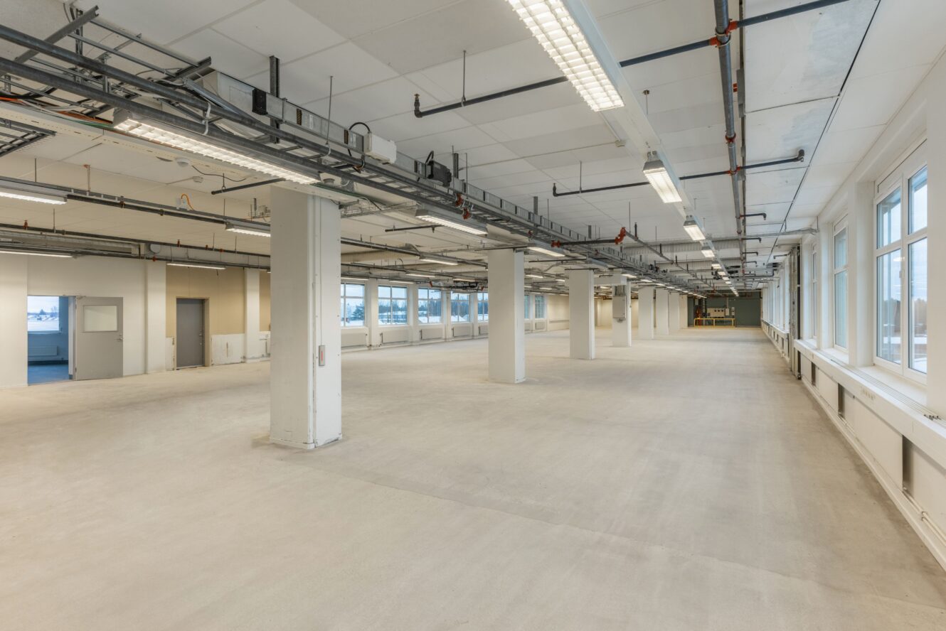 Stor tom industri- eller kontorslokal med betonggolv, synliga rör i taket, stödpelare och fönster längs ena sidan.