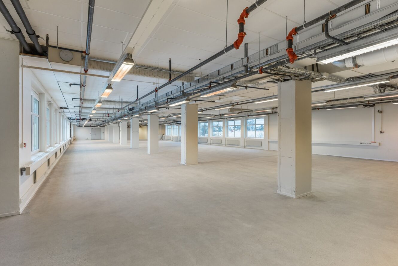 Stor, tom industrilokal med synliga rör i taket, vita pelare och stora fönster längs väggarna. Golvet är kalt och lysrör är monterade i taket.