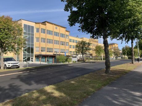 En stor gul byggnad med flera fönster ligger längs en lugn gata som kantas av träd och parkerade bilar en solig dag.