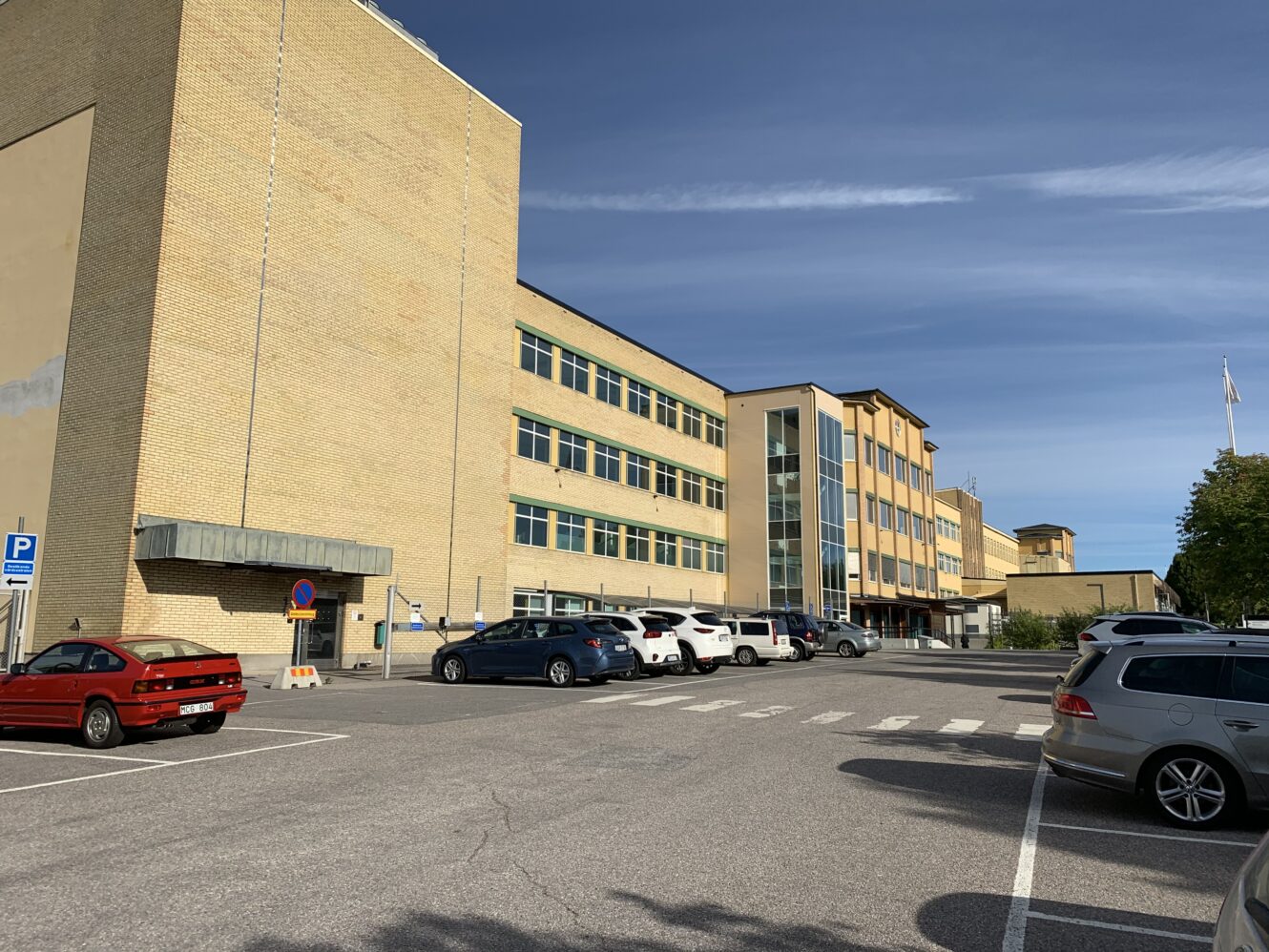 En stor beige tegelbyggnad med flera våningar, bilar parkerade framför och en klarblå himmel ovanför.