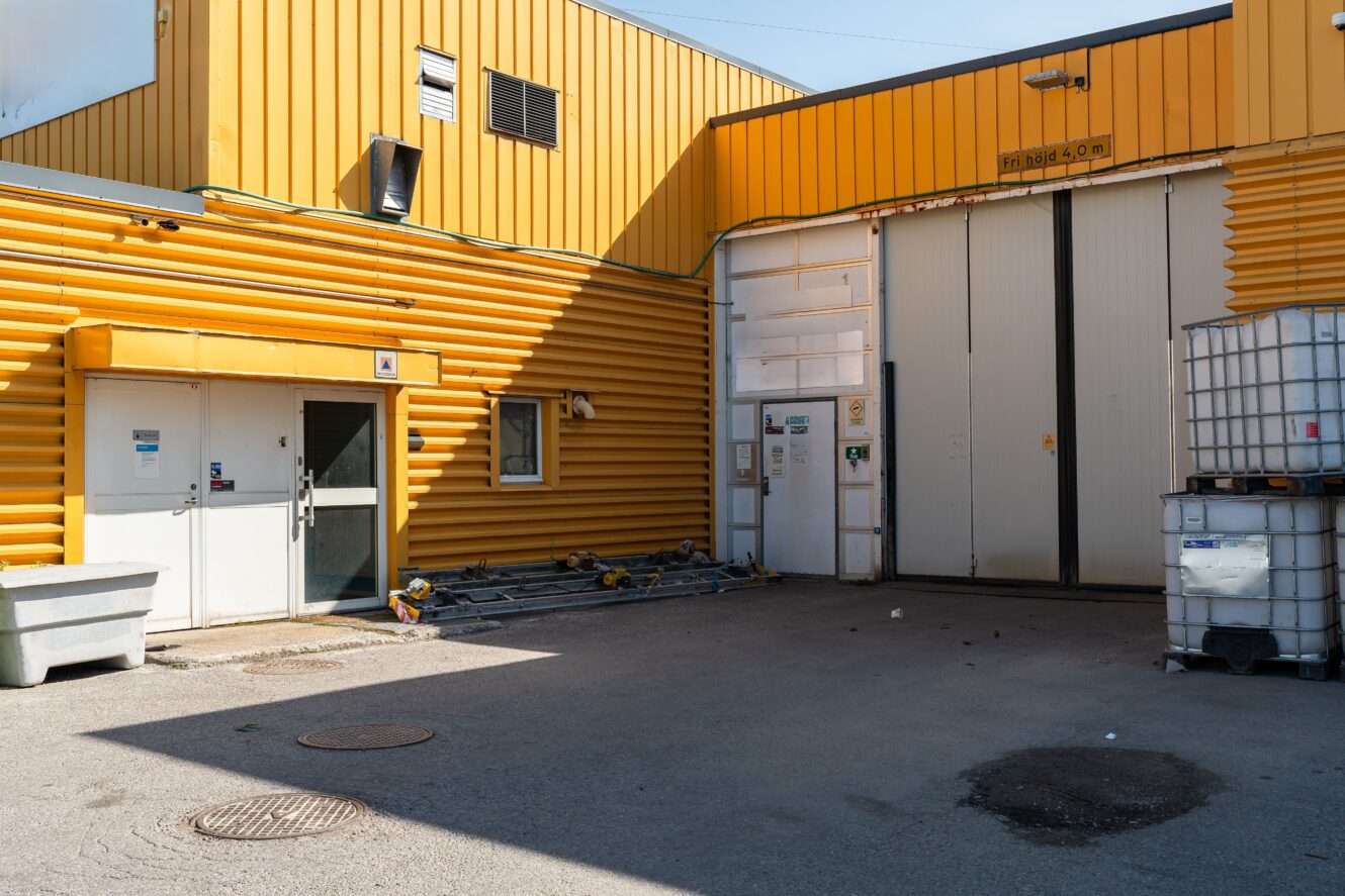 Industrianläggning med gula korrugerade metallväggar, stora vita dörrar, utspridd utrustning och förvaringscontainrar på ett asfalterat utomhusområde.