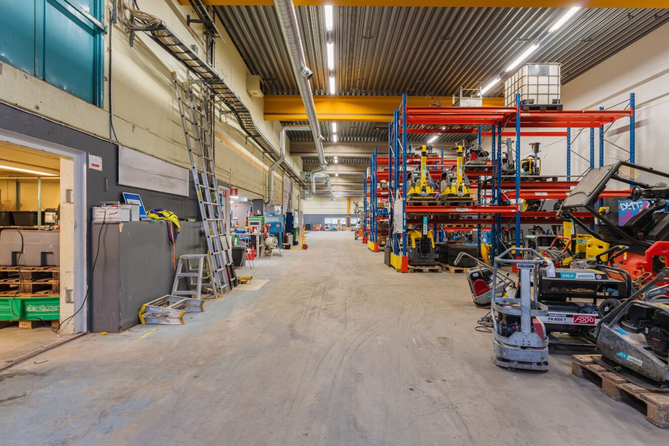 Ett rymligt industrilager med hyllor, maskiner, verktyg och utrustning placerade längs båda sidor av en bred mittgång.