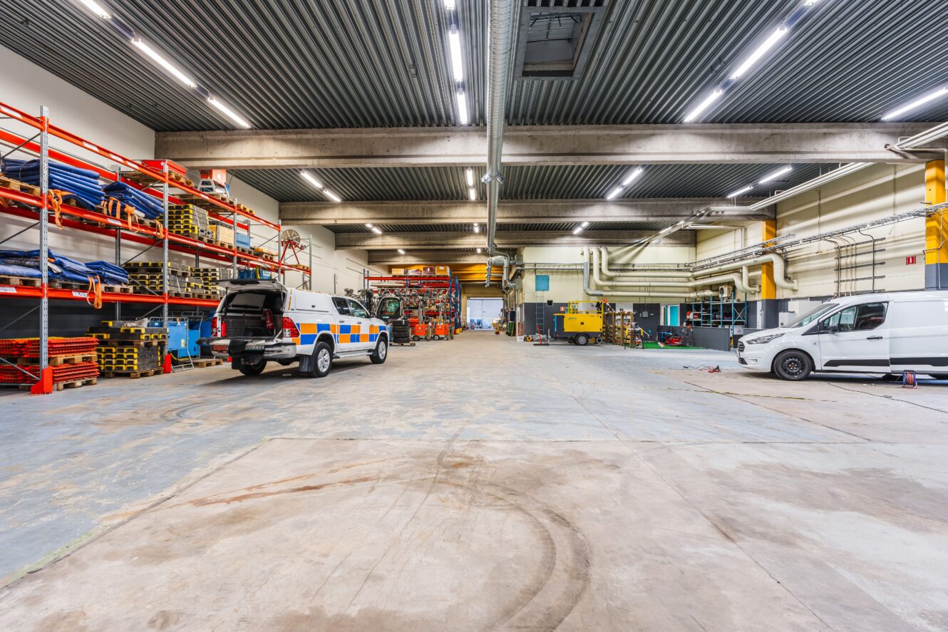 Stort industrigarage med högt i tak, två nyttofordon, metallhyllor, utrustning och olika verktyg organiserade längs väggarna.