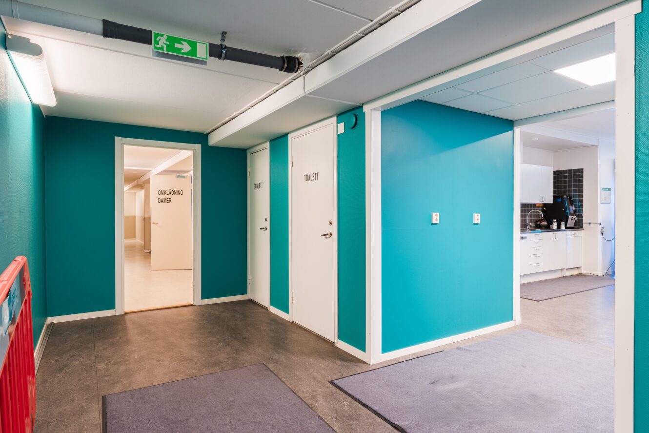 En hall med blågröna väggar, grått golv och flera vita dörrar märkta "Toalett" och "Omklädnings Damer"; en intilliggande öppen yta avslöjar ett pentry.
