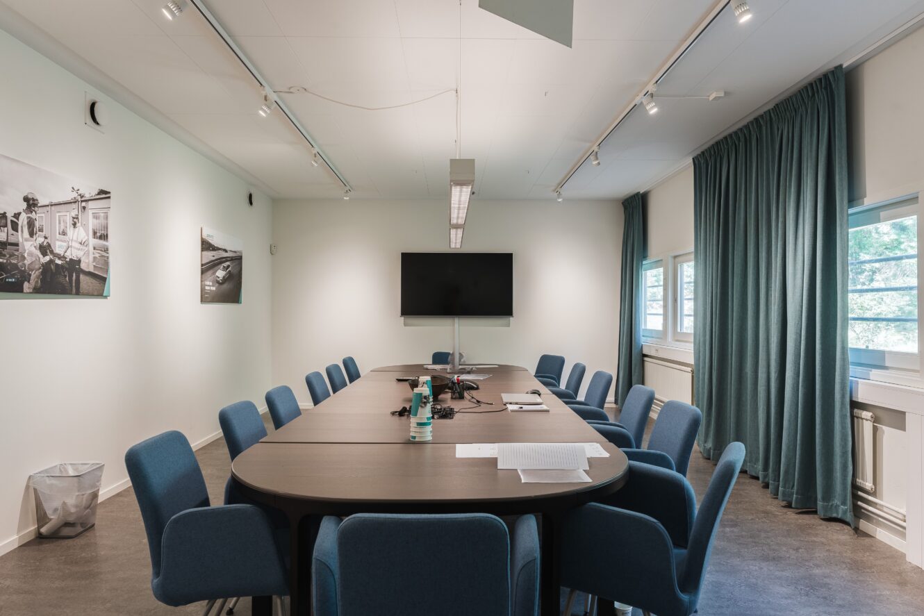 Ett konferensrum med ett långt bord, blå stolar, en väggmonterad TV, några papper och en flaska på bordet samt stora fönster med blågröna gardiner.