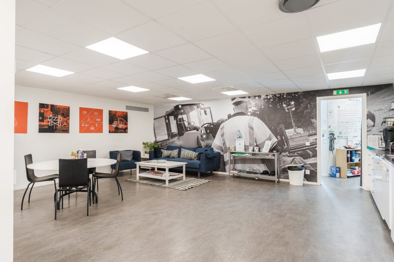 Modernt fikarum med ett runt bord, svarta stolar, blå soffor, väggkonst och en stor väggmålning med byggnadsarbetare; pentryt syns till höger.