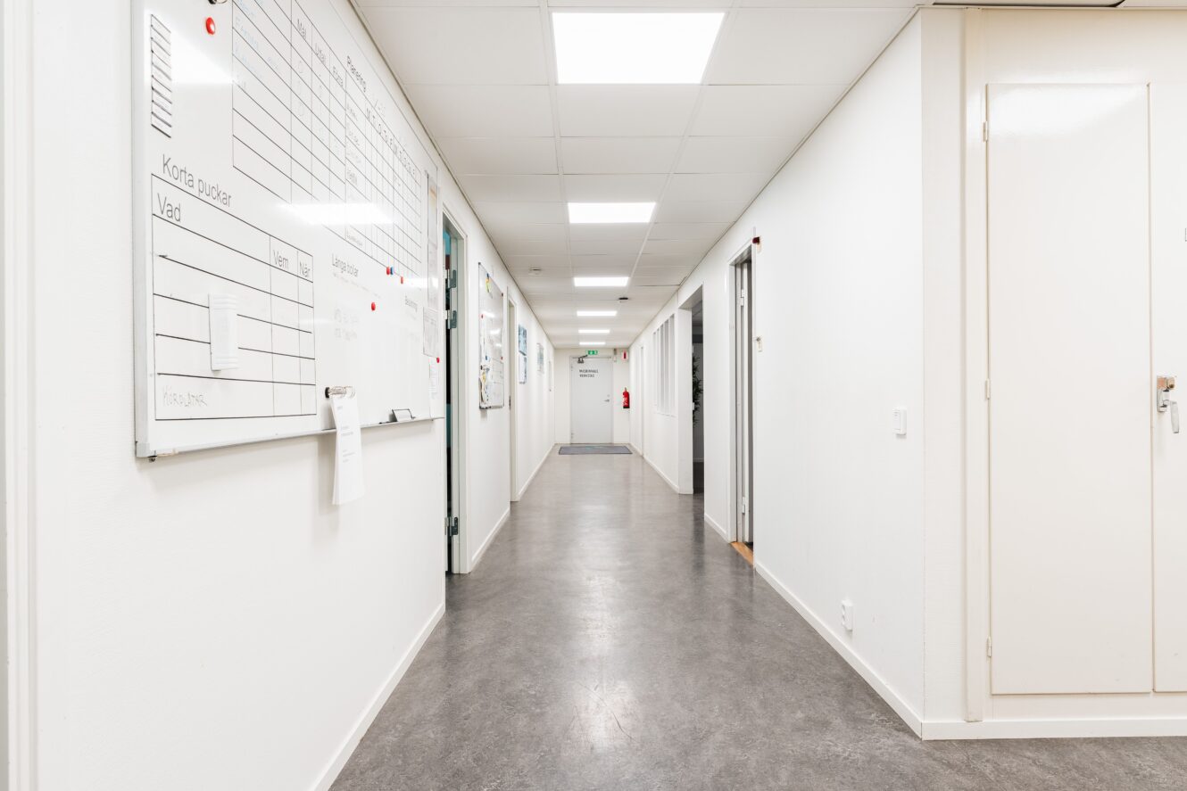 En ren, väl upplyst korridor med vita väggar, en whiteboard till vänster, stängda dörrar på båda sidor och en brandsläckare längst bort.
