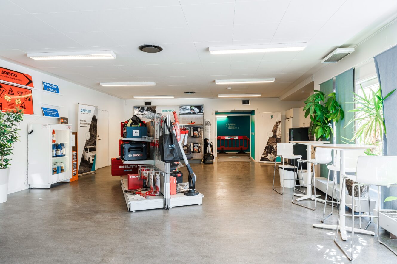 Ett utställningsrum med utställda byggmaskiner och verktyg, vita väggar, stora fönster, krukväxter och höga bord med stolar.