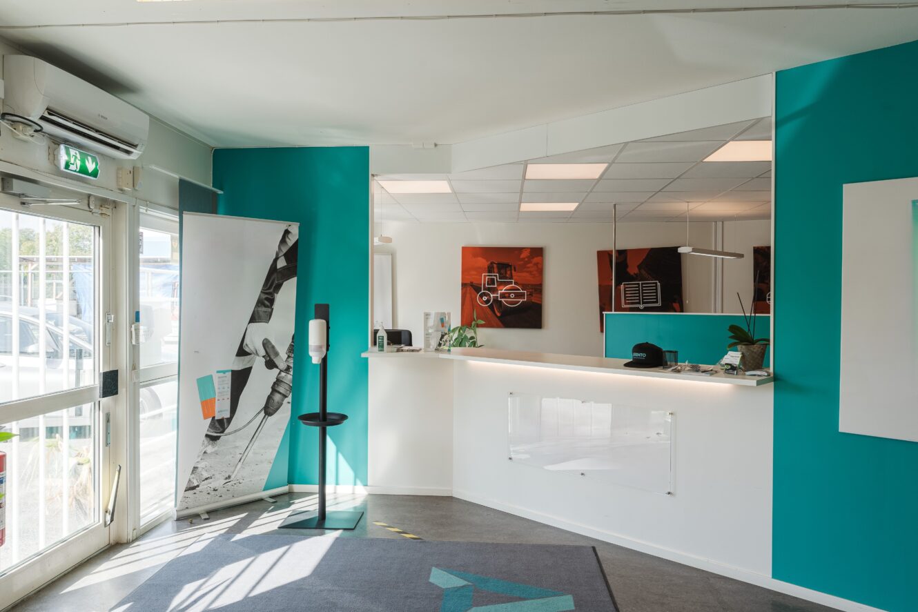 Modern reception med turkosfärgade väggar, en vit disk, konstverk, en desinfektionsstation och stora fönster som släpper in naturligt ljus.