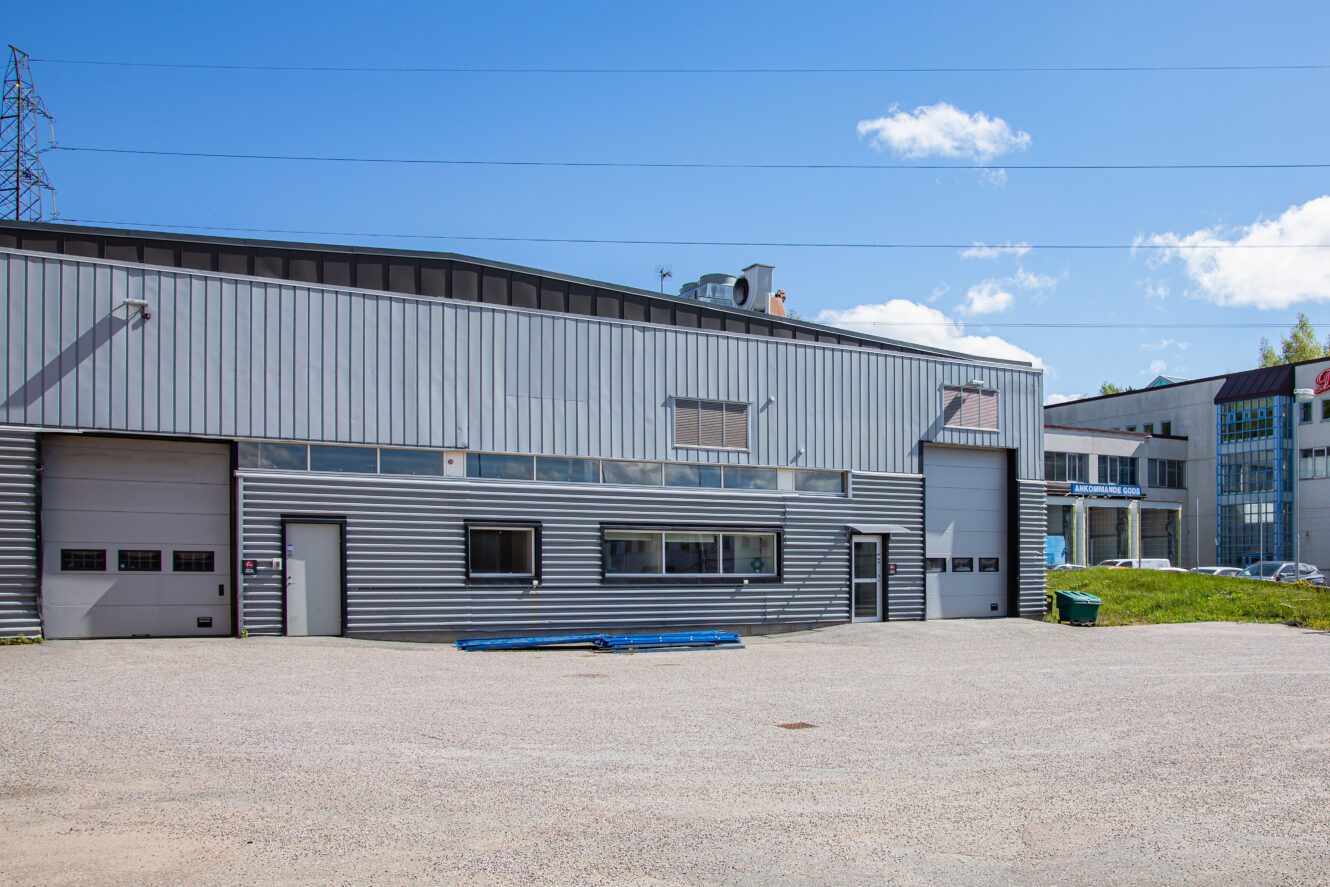 Industriell lagerbyggnad med metallfasad, flera garageportar och en grusad parkeringsplats under en klarblå himmel.