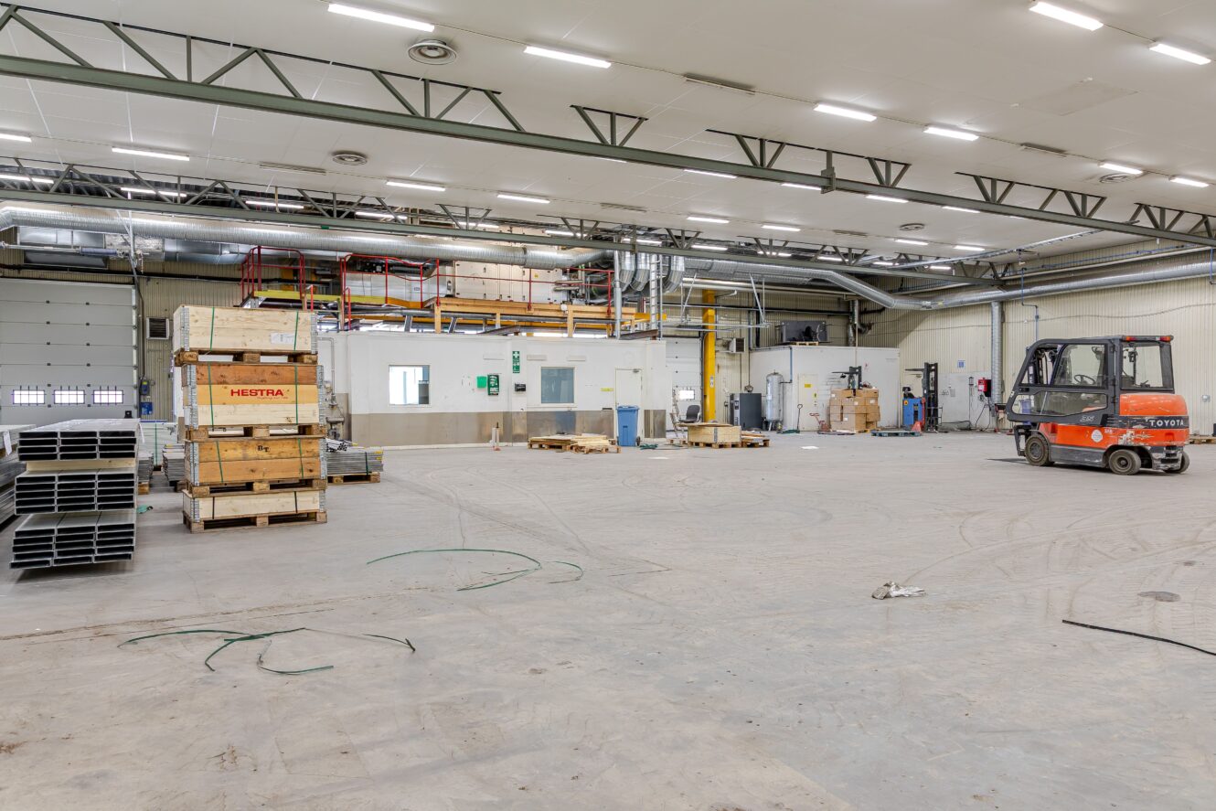 Rymlig industriell lagerlokal med en gaffeltruck, staplade pallar, lådor, hyllor och diverse utrustning på ett betonggolv.