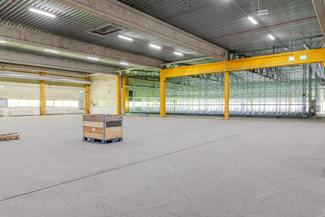 Rymlig, tom lagerlokal med betonggolv, högt i tak, gul traverskran och några pallar placerade nära mitten.