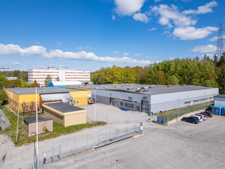 En inhägnad industrianläggning med flera lagerbyggnader, intilliggande parkering och omgivande träd under en delvis molnig himmel.