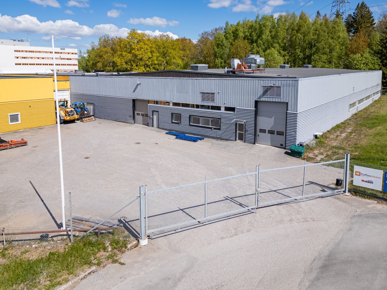 En inhägnad industribyggnad med grå fasad och flera garageportar, omgiven av träd och en annan gul byggnad under en blå himmel.