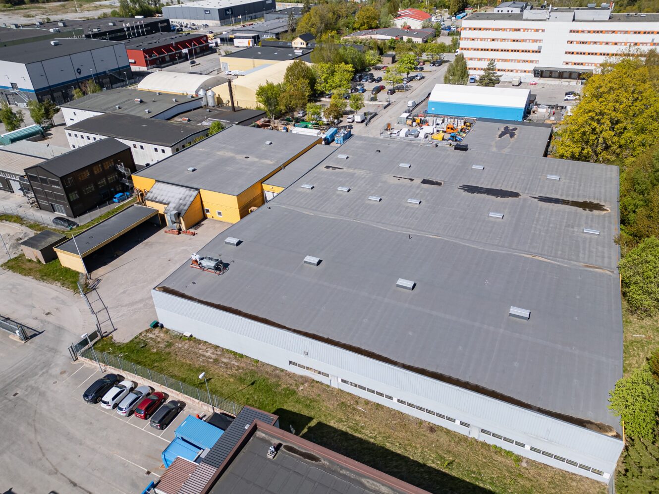 Flygbild över ett industriområde med flera stora lagerbyggnader, parkerade bilar, träd och omgivande vägar en klar dag.