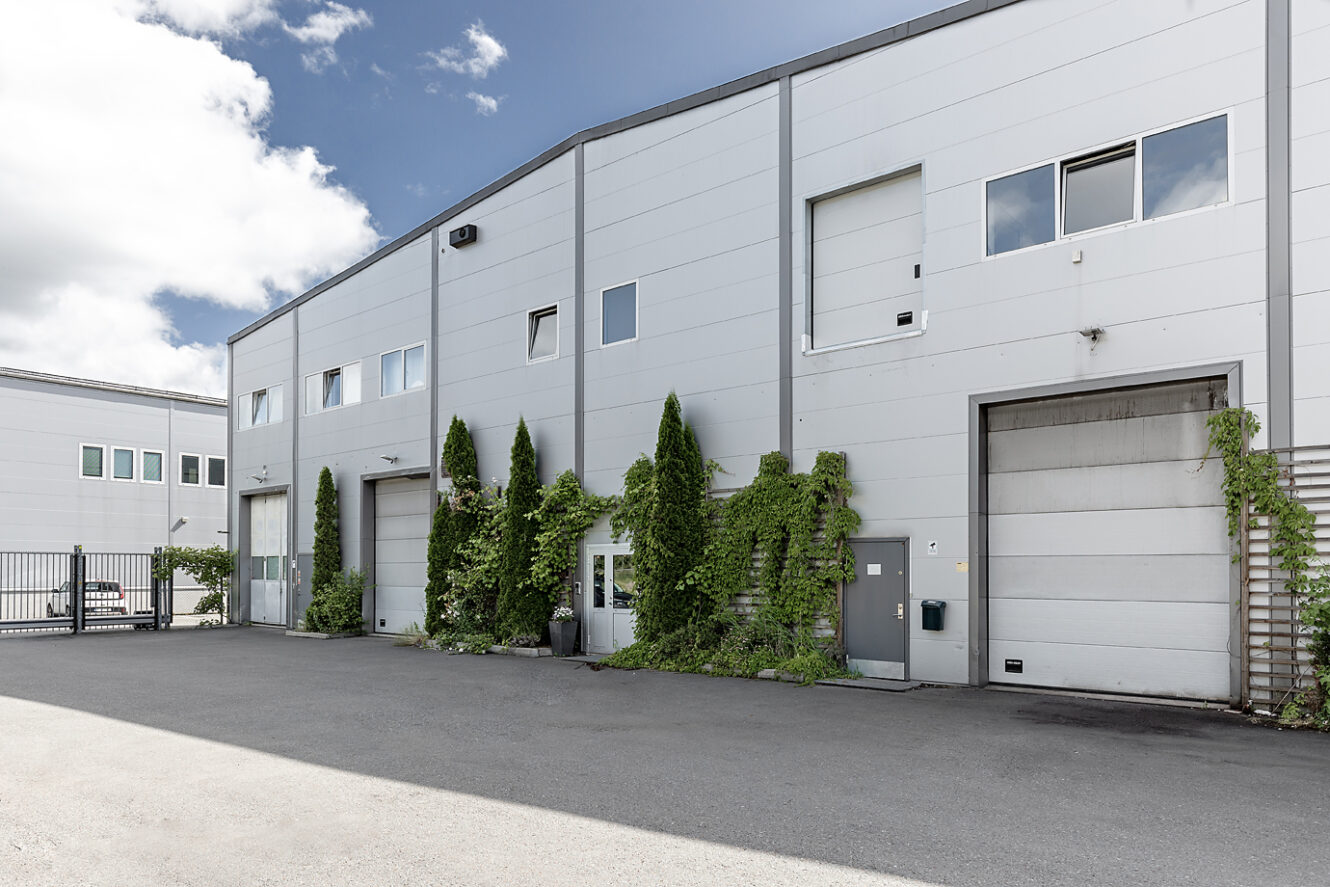 En modern industribyggnad med vit fasad, flera garageportar, fönster och gröna vinrankor som växer längs ytterväggen.