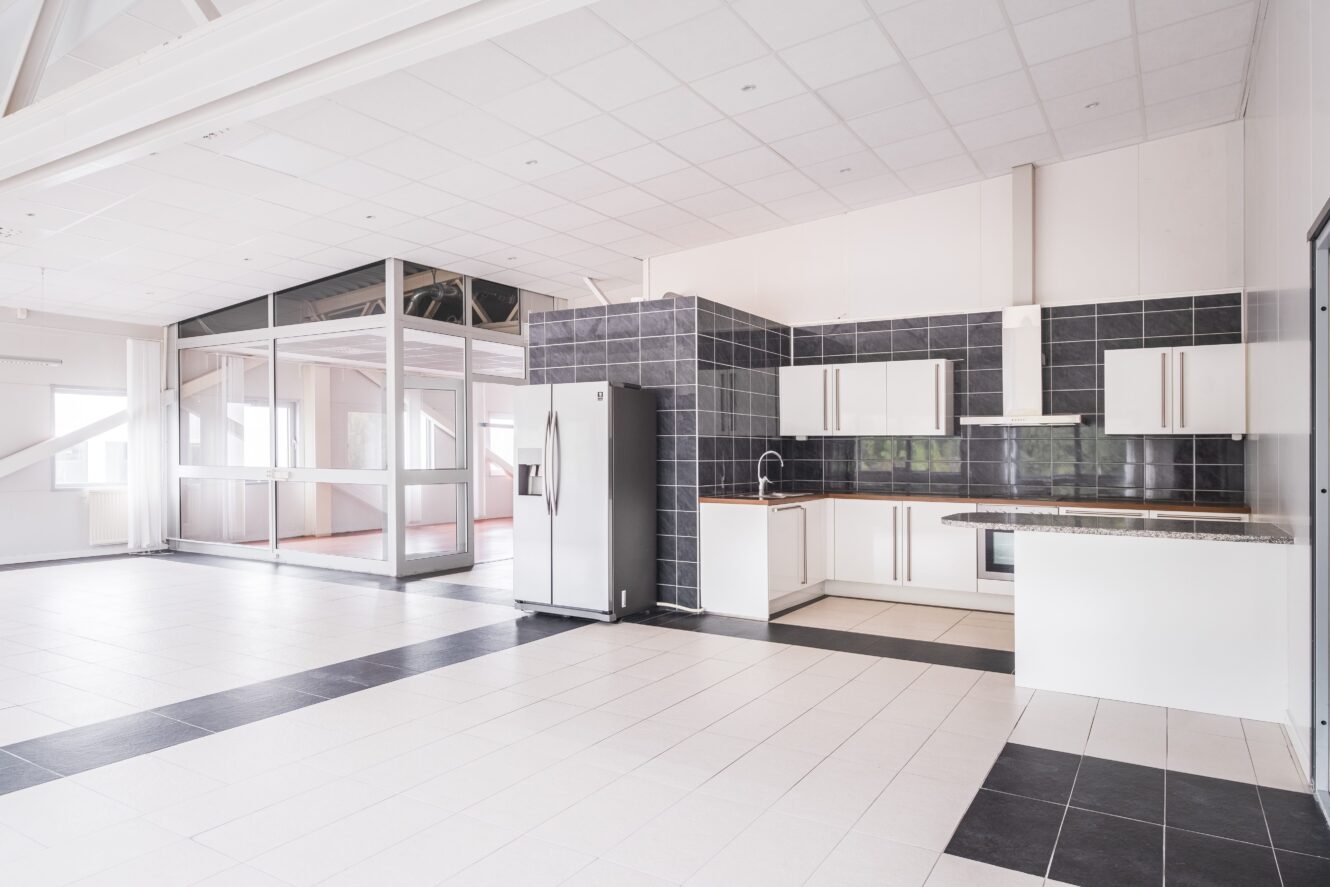 Rymligt modernt kök med vita skåp, kylskåp i rostfritt stål, svart kaklat stänkskydd och ett angränsande rum med glasväggar.