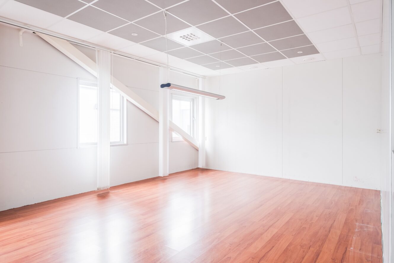 Ett tomt rum med ljusa väggar, stora fönster, en hängande armatur och ett golv i trästil.