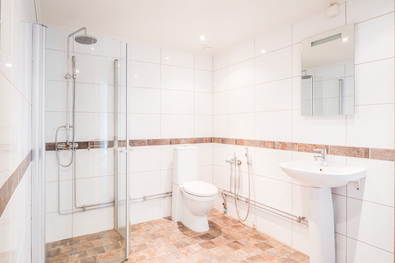 Ett modernt badrum med dusch i glas, toalett, väggmonterat handfat, stor spegel och vitkaklade väggar med brunt mönstrat golvklinker.