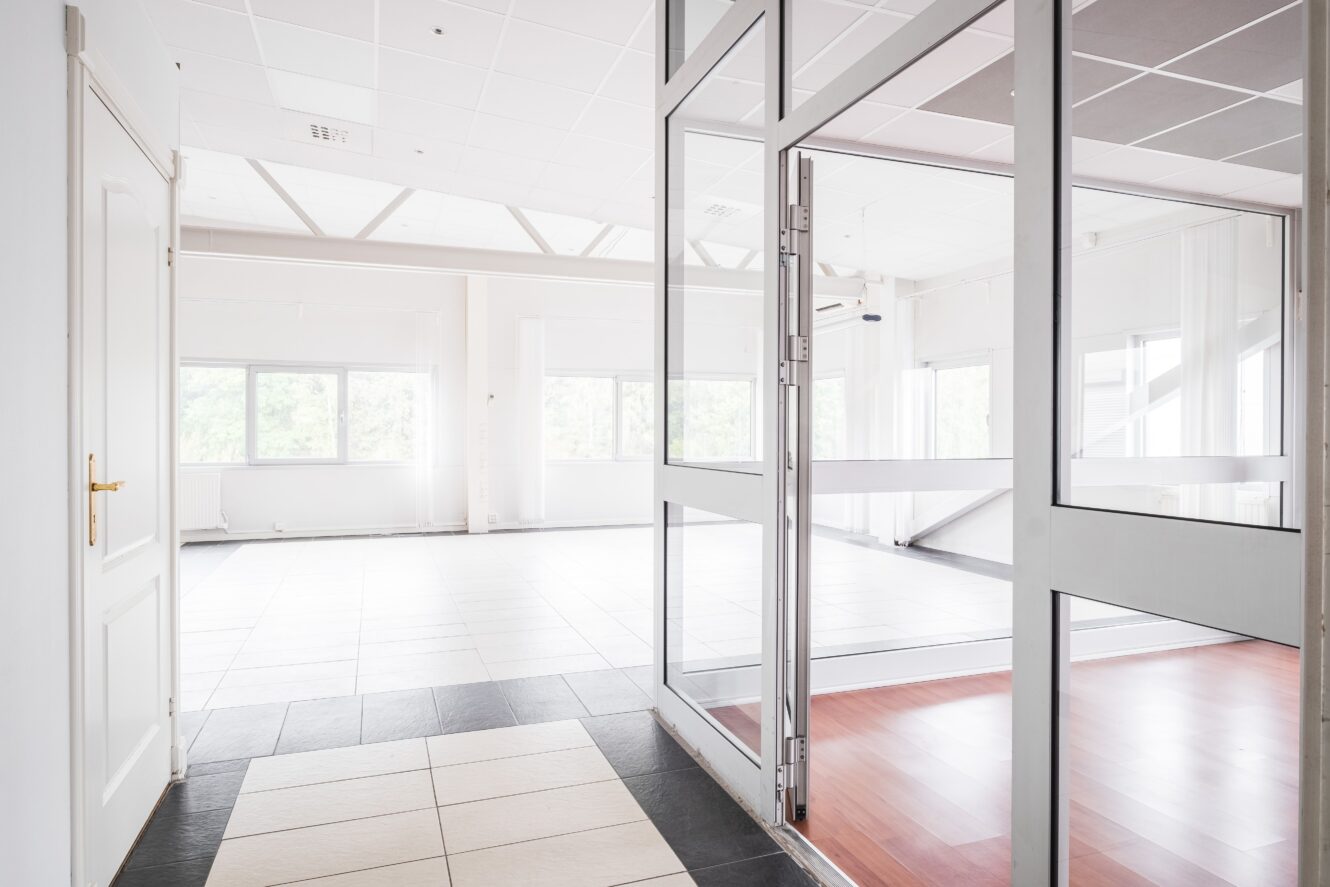 Ett ljust, tomt rum med stora fönster, vita väggar och en glasdörr som skiljer klinkergolv från ett utrymme med trälaminatgolv.