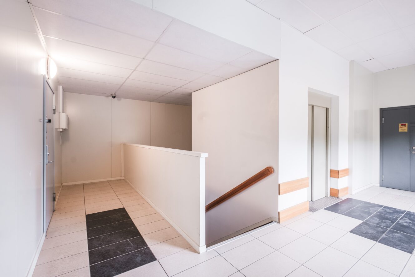 En ljus hall med klinkergolv, vita väggar, en hiss, en grå dörr och en ledstång som leder nerför en kort trappa.