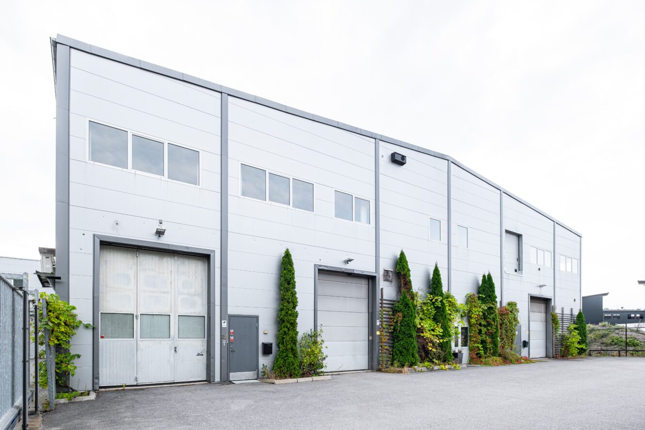 En modern industriell lagerbyggnad med grå metallfasad, flera garageportar, fönster och gröna klätterväxter längs fasaden.