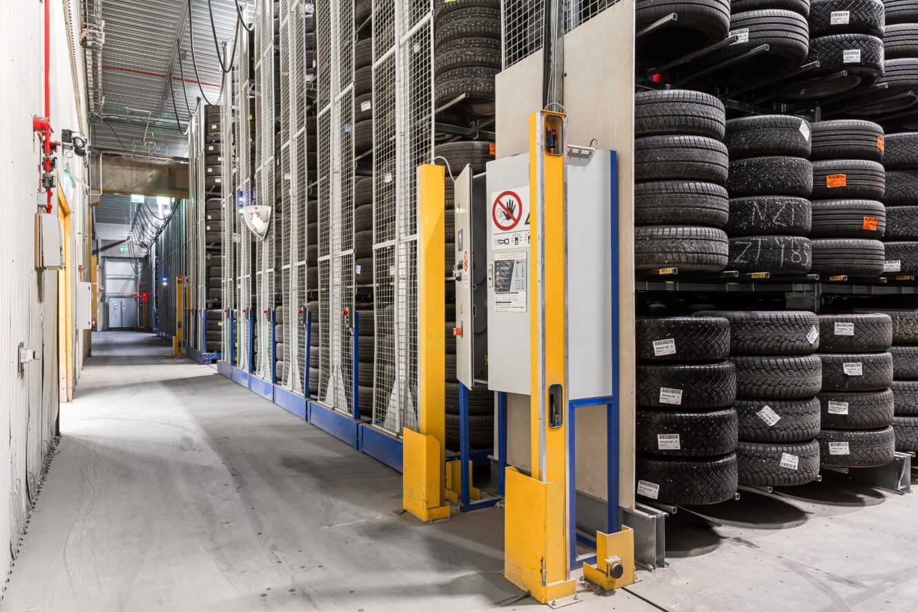 En lagergång med höga ställningar fyllda med staplade däck och en kontrollpanel med säkerhetsskyltar och stark belysning.