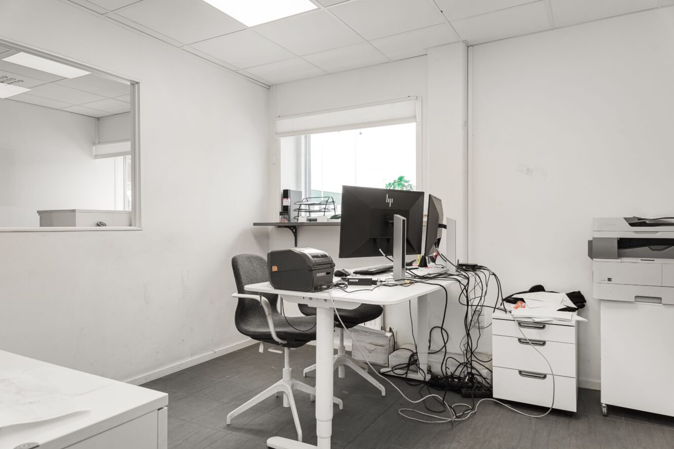 Ett litet, prydligt kontor med skrivbord, dubbla skärmar, kontorsstol, skrivare och spridda papper, med vita väggar och ett fönster som ger naturligt ljus.