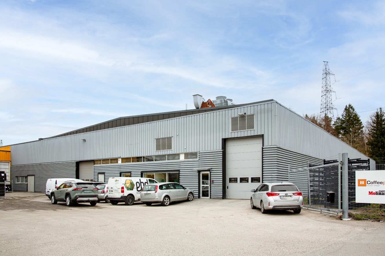 En stor grå industrilagerbyggnad med flera fordon parkerade utanför, inklusive en vit skåpbil med sidodörren öppen, på en asfalterad tomt under en delvis molnig himmel.