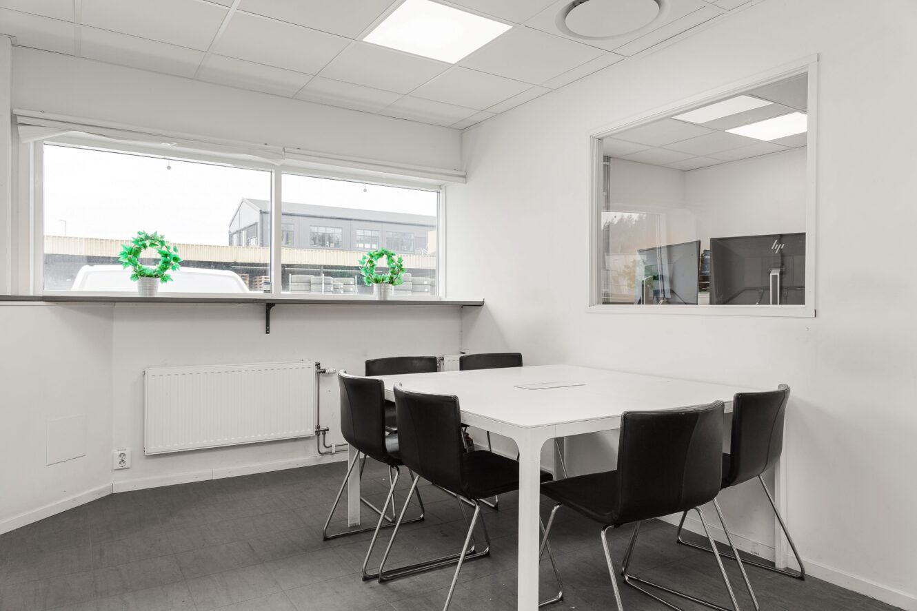 Ett litet, modernt mötesrum med ett vitt bord, sex svarta stolar, två fönster och minimal inredning, inklusive två gröna växter på fönsterbrädan.