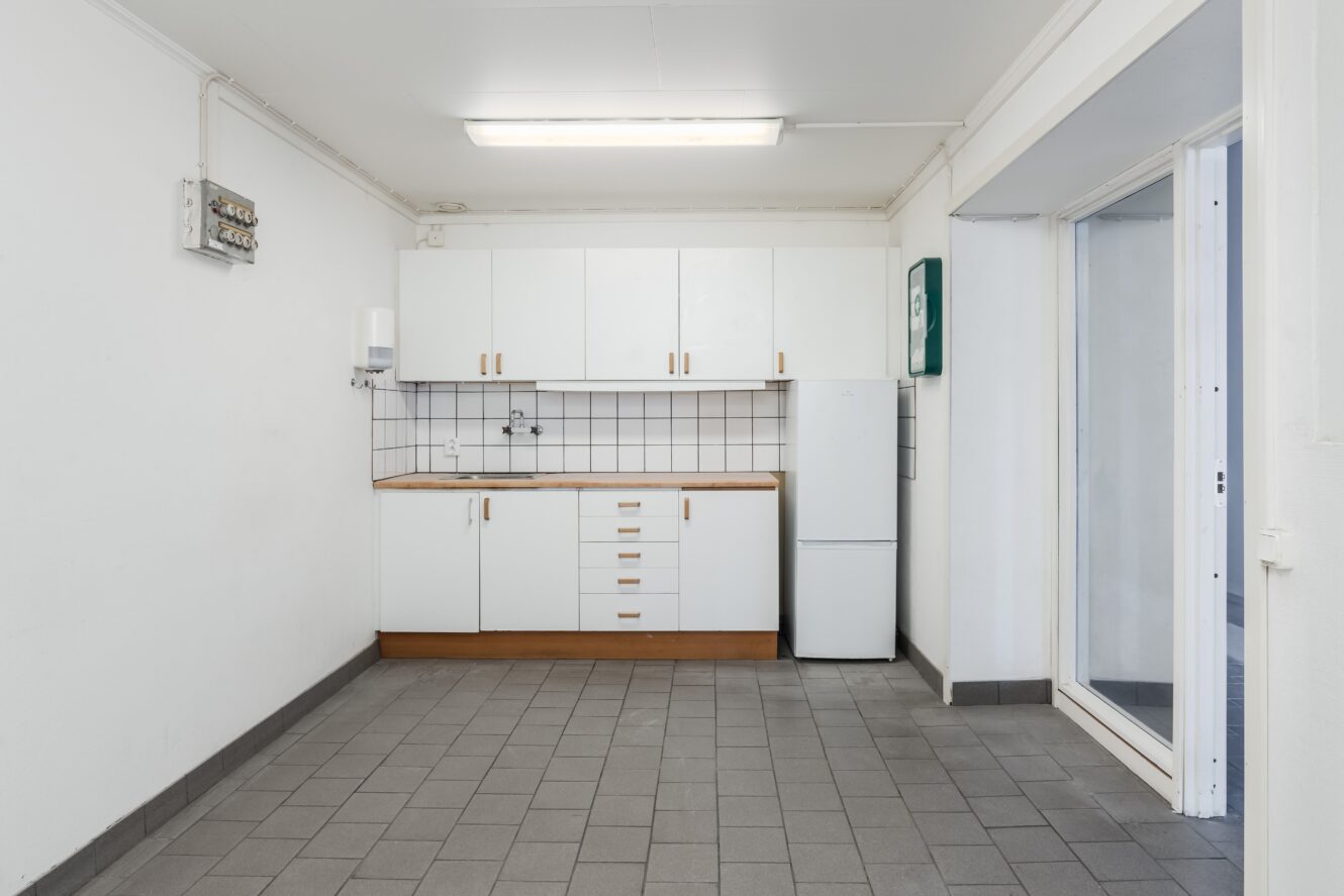 Ett litet, tomt pentry med vita skåp, kaklat stänkskydd, kylskåp och grått klinkergolv.