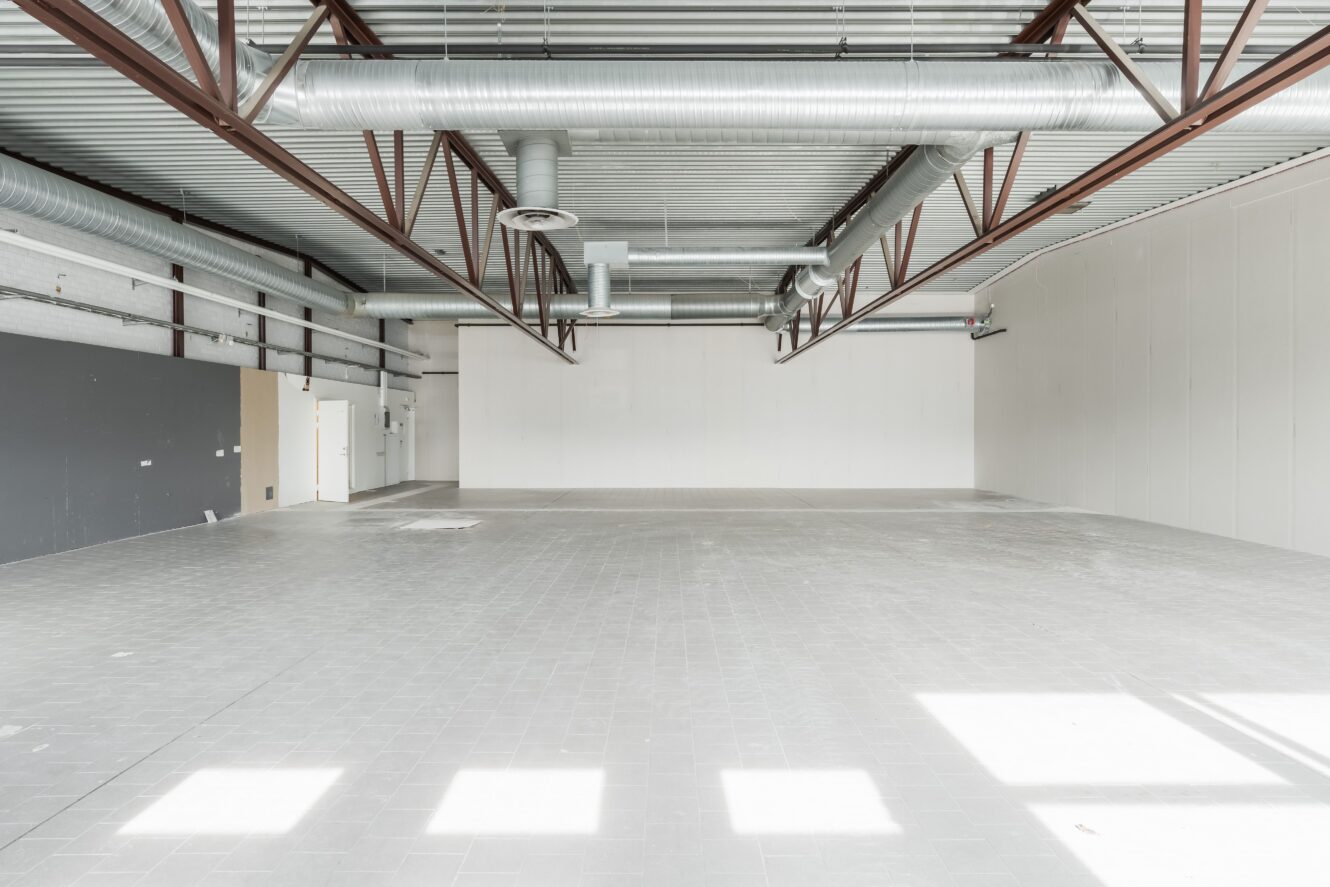 Tomt industrilokal med klinkergolv, takbjälkar i synlig metall, ventilationskanaler, vita väggar och starkt naturligt ljus från osynliga fönster.