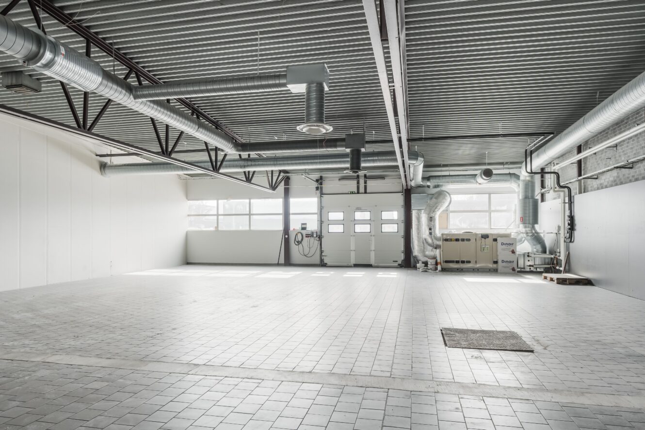 En tom industrilokal med klinkergolv, stora takventiler, vita väggar och en upprullbar garageport som släpper in naturligt ljus.