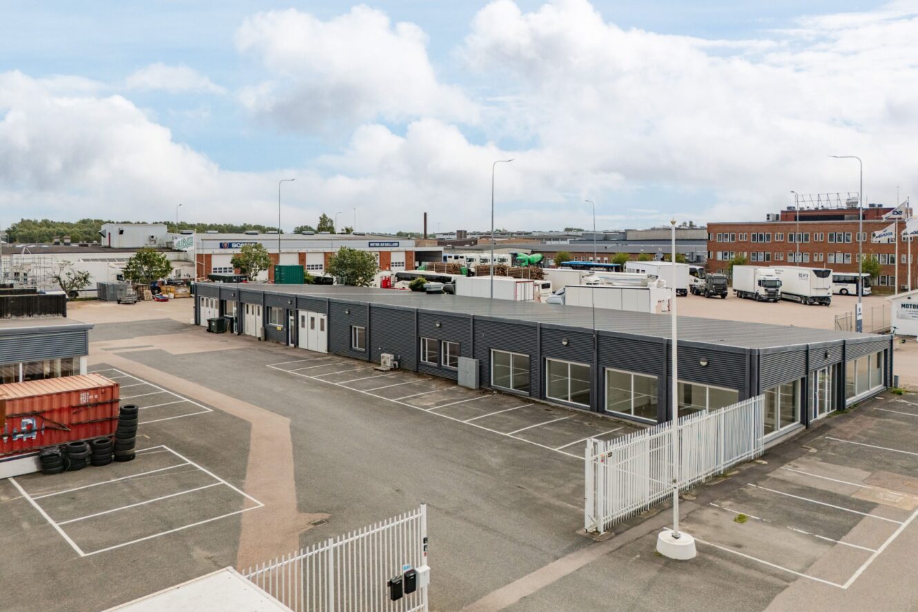 En stor industritomt med en grå envåningsbyggnad, flera parkerade lastbilar, fraktcontainrar och tomma parkeringsplatser under en delvis molnig himmel.