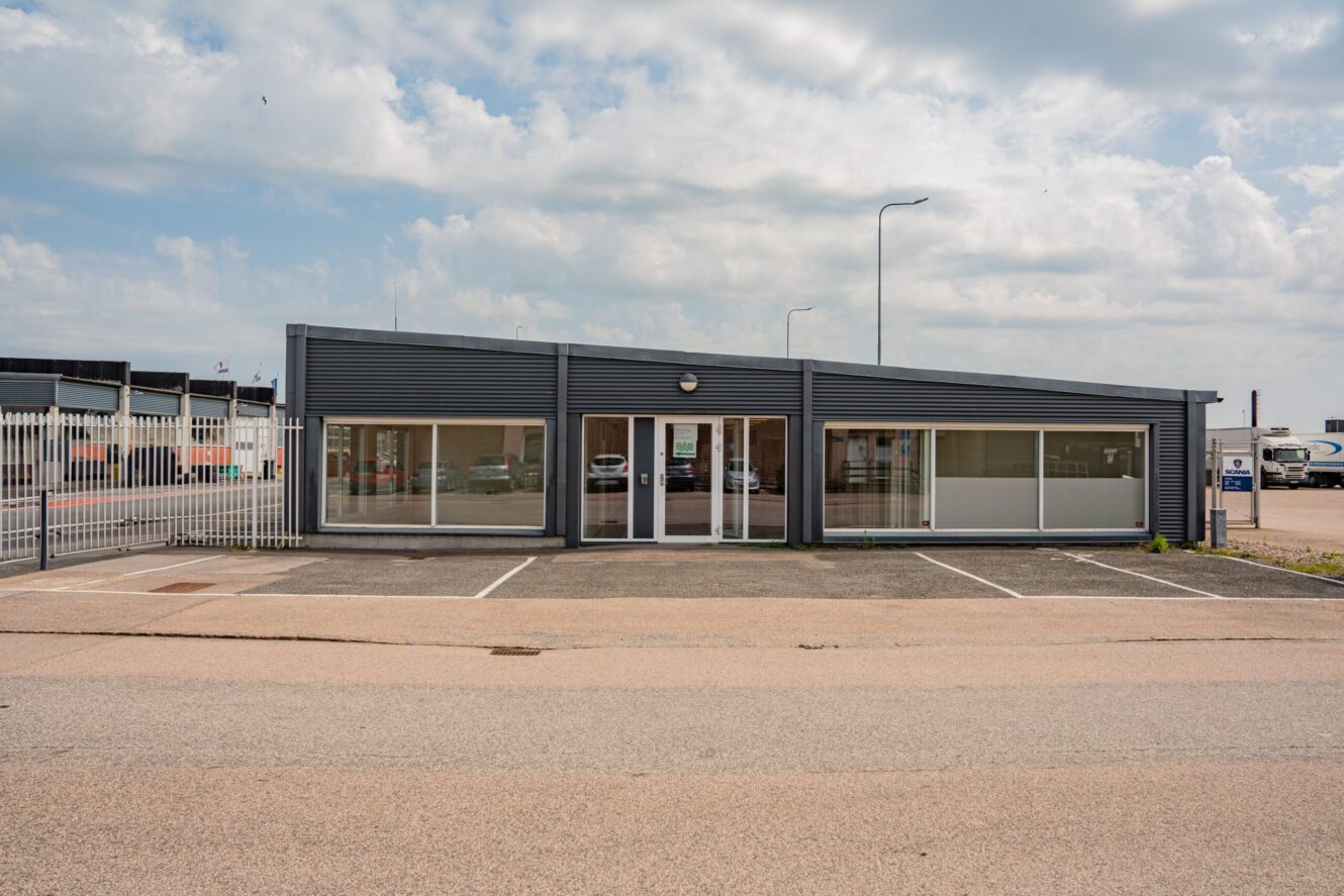 En grå affärsbyggnad i en våning med stora fönster och tomma parkeringsplatser framför, under en delvis molnig himmel.